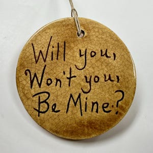 May include: A round, ceramic ornament with a crackle glaze finish. The text "Will you, Won't you, Be Mine?" is written in black script. A natural twine loop is attached for hanging.