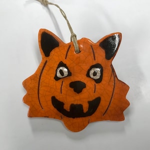 May include: Orange ceramic Halloween decoration shaped like a jack-o'-lantern cat face with black accents. The cat's eyes are silver, and the mouth is a carved-out grin. A twine loop is attached for hanging. The decoration has a cracked, textured surface.