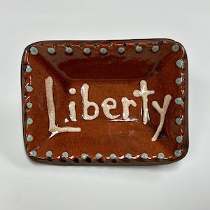 May include: A rectangular ceramic tray with a glossy brown glaze and a crackle finish. The word Liberty is written in raised, off-white, crackled lettering in the center. The edges are decorated with a border of small, raised, light blue dots.