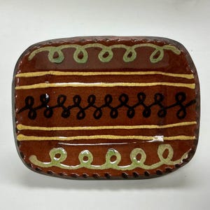 May include: A rectangular ceramic dish with a brown base and decorative elements. The dish features horizontal yellow and black lines, with green and black swirl patterns. The edges are dark brown, and the glaze has a crackled appearance.