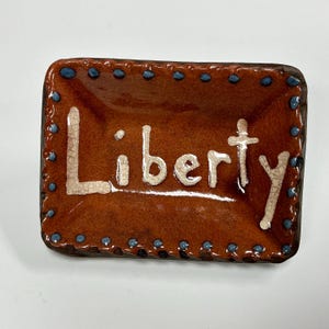 May include: A rectangular ceramic tray with a glossy brown glaze and a crackled white finish on the raised lettering that spells out the word Liberty. The edges are decorated with a border of small, raised blue dots along the entire perimeter.