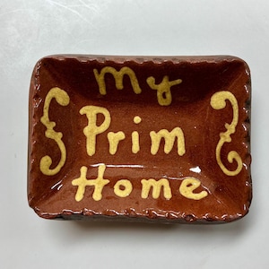 May include: A rectangular, brown ceramic dish with the words "my Prim Home" in yellow lettering. The dish has a textured, scalloped edge and decorative yellow flourishes on the sides.
