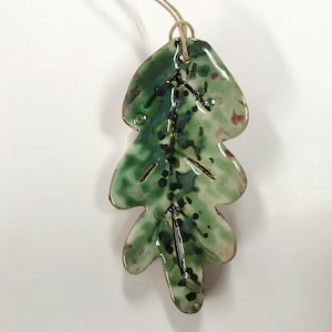 May include: A ceramic leaf ornament with a naturalistic design. The leaf is primarily green with darker green and black accents, and a touch of reddish-brown. It has a glossy finish and a twine loop for hanging.