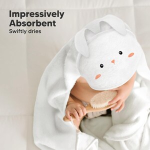 May include: White hooded baby towel with bunny ears and a cartoon face. The towel has black eyes and peach-colored cheeks. The text "Impressively Absorbent" and "Swiftly dries" is in the upper left corner. A baby is holding a wooden brush.