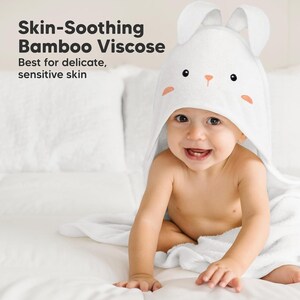 May include: A smiling baby wearing a white hooded towel with bunny ears. The towel has a cute bunny face design with black eyes and peach accents. Text on the left reads "Skin-Soothing Bamboo Viscose" and "Best for delicate, sensitive skin."