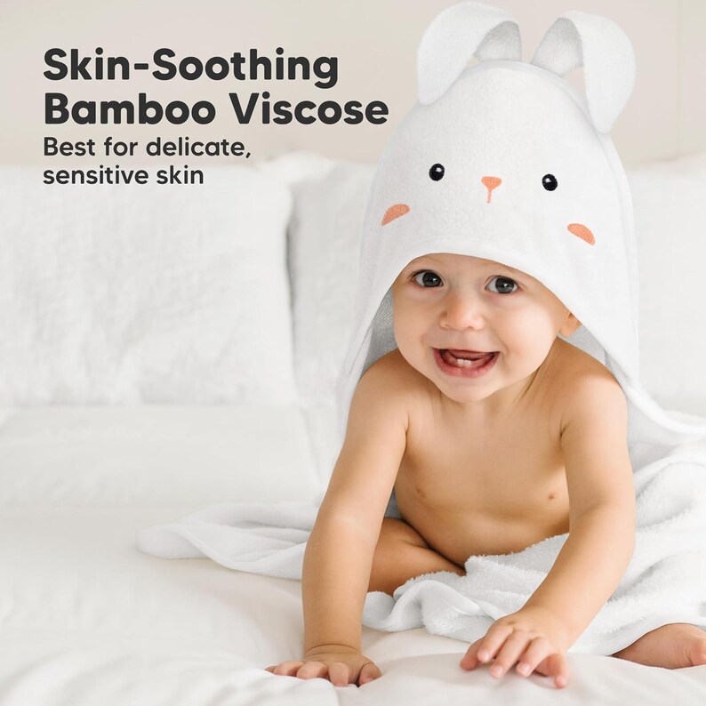 May include: A smiling baby wearing a white hooded towel with bunny ears. The towel has a cute bunny face design with black eyes and peach accents. Text on the left reads "Skin-Soothing Bamboo Viscose" and "Best for delicate, sensitive skin."