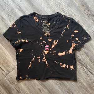 May include: A black cropped t-shirt with a bleach tie-dye pattern and a red lips patch on the front.
