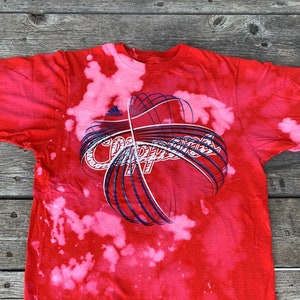 May include: Red t-shirt with a white bleach tie-dye pattern. The shirt has a blue and white graphic of the word "Clippers" with a basketball design.