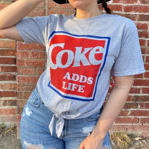 May include: A gray t-shirt with a red and blue graphic that says "Coke Adds Life".