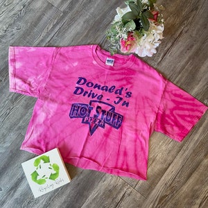 May include: Pink tie-dye cropped t-shirt with a purple and white logo that says "Donald's Drive-In Hot Stuff Pizza".