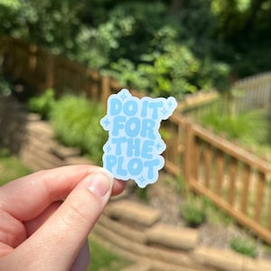May include: A light blue sticker with the phrase "DO IT FOR THE PLOT" in a bubble font. The sticker has a unique shape with star accents and is held in a hand against a blurred outdoor background.