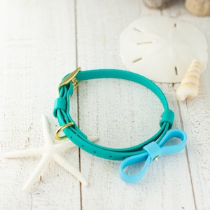 May include: Teal blue adjustable pet collar with a light blue bow. The collar is made of a smooth, rubber-like material.  The gold-toned buckle is visible. This stylish pet accessory is perfect for small dogs.