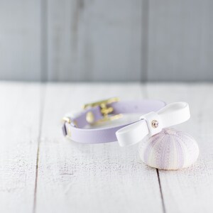 May include: A lavender leather pet collar with a white bow. The bow has a small rhinestone accent. The collar is shown on a white wooden surface next to a lavender sea urchin. The collar is a stylish accessory for small pets.