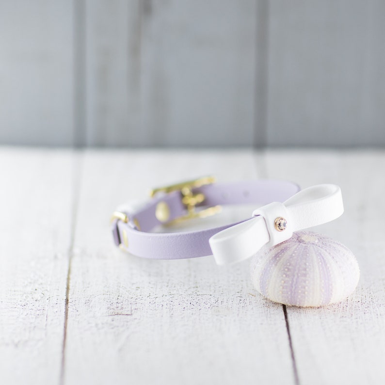 May include: A lavender leather pet collar with a white bow. The bow has a small rhinestone accent. The collar is shown on a white wooden surface next to a lavender sea urchin. The collar is a stylish accessory for small pets.
