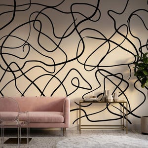 May include: A modern living room with a light pink sofa, clear acrylic chair, and a gold side table. The wall features a large abstract black line design. A white fluffy rug and a potted plant complete the decor.