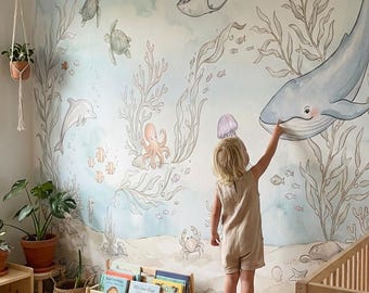 May include: A child's bedroom with a whimsical ocean-themed mural. The mural features a whale, sea turtles, and other marine life. A child stands on a stool, pointing at the whale. The room includes a crib, bookshelf, and a cat.
