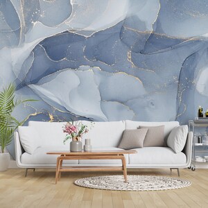 May include: A white sofa with pillows sits in front of a wall with a blue and gold abstract pattern. The sofa is in a living room setting with a wooden coffee table and a plant in a white pot.