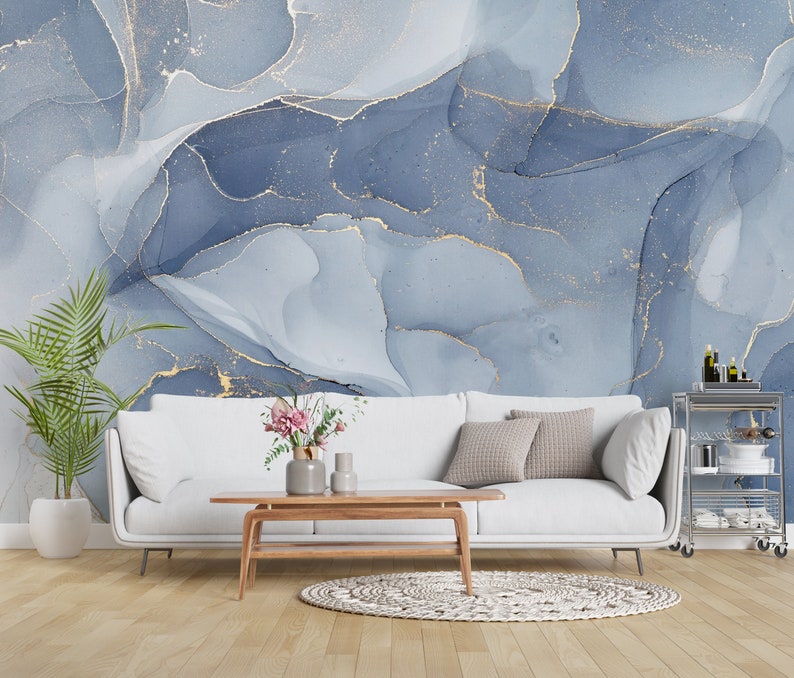 May include: A white sofa with pillows sits in front of a wall with a blue and gold abstract pattern. The sofa is in a living room setting with a wooden coffee table and a plant in a white pot.