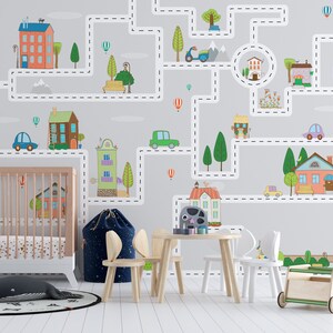 May include: A nursery room with a gray wall mural depicting a city with houses, cars, trees, and a dashed line road. The mural is a fun and colorful design for a child's room. There is a crib, a wooden table and chairs, and a toy wagon in the room.