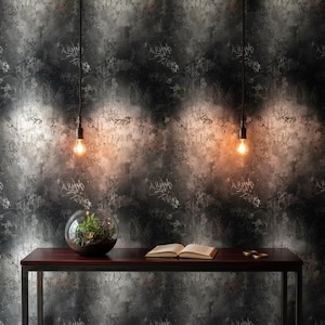 May include: A dark wood table with a metal frame supports a glass terrarium, an open book, and small gold coins. Two hanging light bulbs illuminate the scene against a textured, graffiti-style wall. The overall aesthetic is modern and industrial.