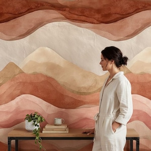 Pode incluir: Uma mulher de macacão branco está perto de uma mesa de madeira com uma estrutura de metal preta. O fundo apresenta um mural em estilo aquarela de cadeias de montanhas em camadas e tons terrosos. Uma pequena planta e uma caneca estão sobre a mesa.