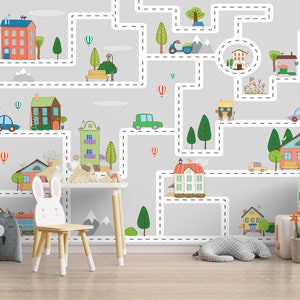 May include: A children's room with a gray wall featuring a wallpaper with a city scene. The wallpaper has a white dotted line path that goes through the city. The city has houses, trees, cars, and hot air balloons. The room has a white table with a white chair, a stuffed animal, a gray storage bin with a panda face, and a yellow block with the letter 'B' on it. There is also a potted plant with green leaves and a yellow mug on a wooden stool.