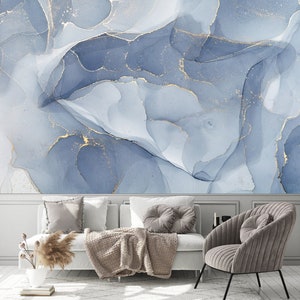 May include: A living room with a white couch and a gray armchair. The wall behind the couch is decorated with a blue and gold abstract painting. The couch has a brown throw blanket and a few pillows. The armchair has a gray pillow.