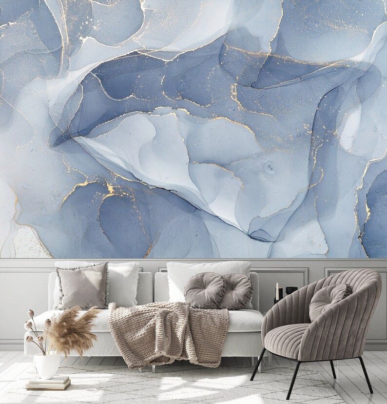 May include: A living room with a white couch and a gray armchair. The wall behind the couch is decorated with a blue and gold abstract painting. The couch has a brown throw blanket and a few pillows. The armchair has a gray pillow.