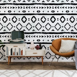 Pode incluir: Um papel de parede com padrão geométrico preto e branco com um toca-discos, uma mesa auxiliar, uma cadeira e uma planta em um vaso. A cadeira é marrom e tem uma almofada branca. A mesa auxiliar é branca e tem um vaso e uma escultura em cima. A planta é verde e tem folhas longas.