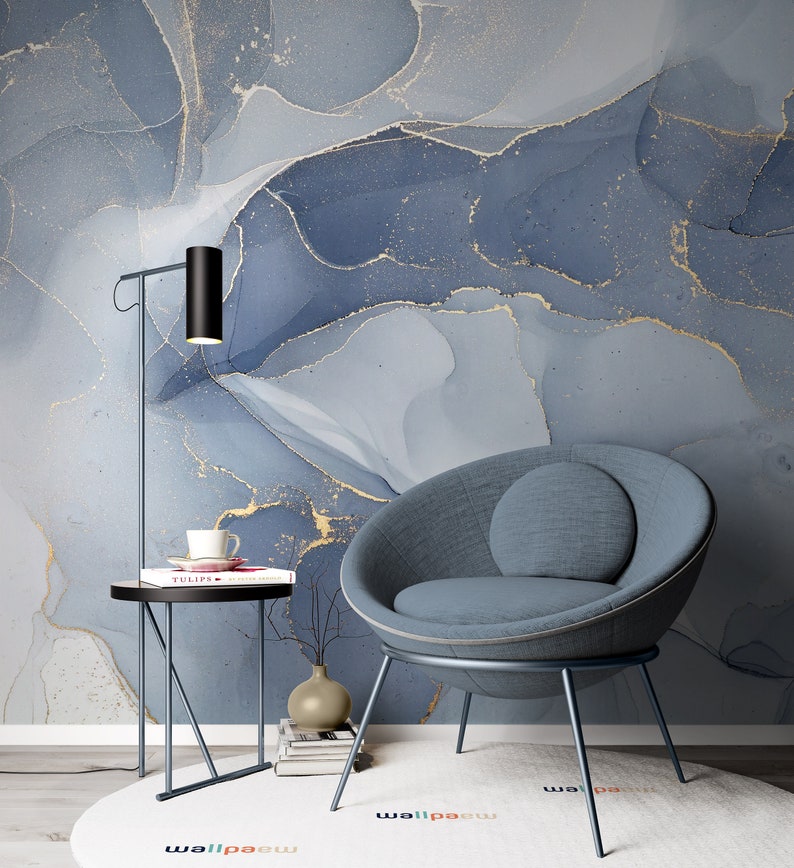 May include: A blue and gold abstract marble wallpaper with a blue armchair and a black side table with a book and a cup on top. The side table has a black metal frame and a round top. The armchair has a round back and a gray fabric seat. The wallpaper has a modern and abstract design.