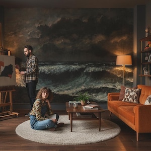 Könnte beinhalten: Innenaufnahme eines Künstlerateliers mit einem Paar. Ein großes Gemälde eines stürmischen Meeres hängt an der Wand. Ein orangefarbenes Sofa, ein Holztisch und ein Bücherregal sind ebenfalls zu sehen. Der Raum hat eine warme, einladende Atmosphäre.