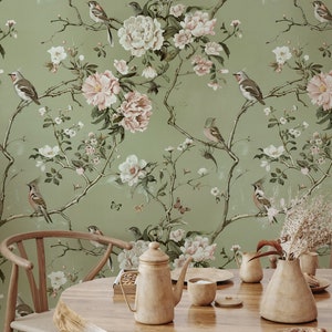 Op de afbeelding: Een groene behang met een bloemenpatroon met roze en witte bloemen, groene bladeren en vogels. Het behang bevindt zich in een eetkameromgeving met een houten tafel, stoelen en een theepot.