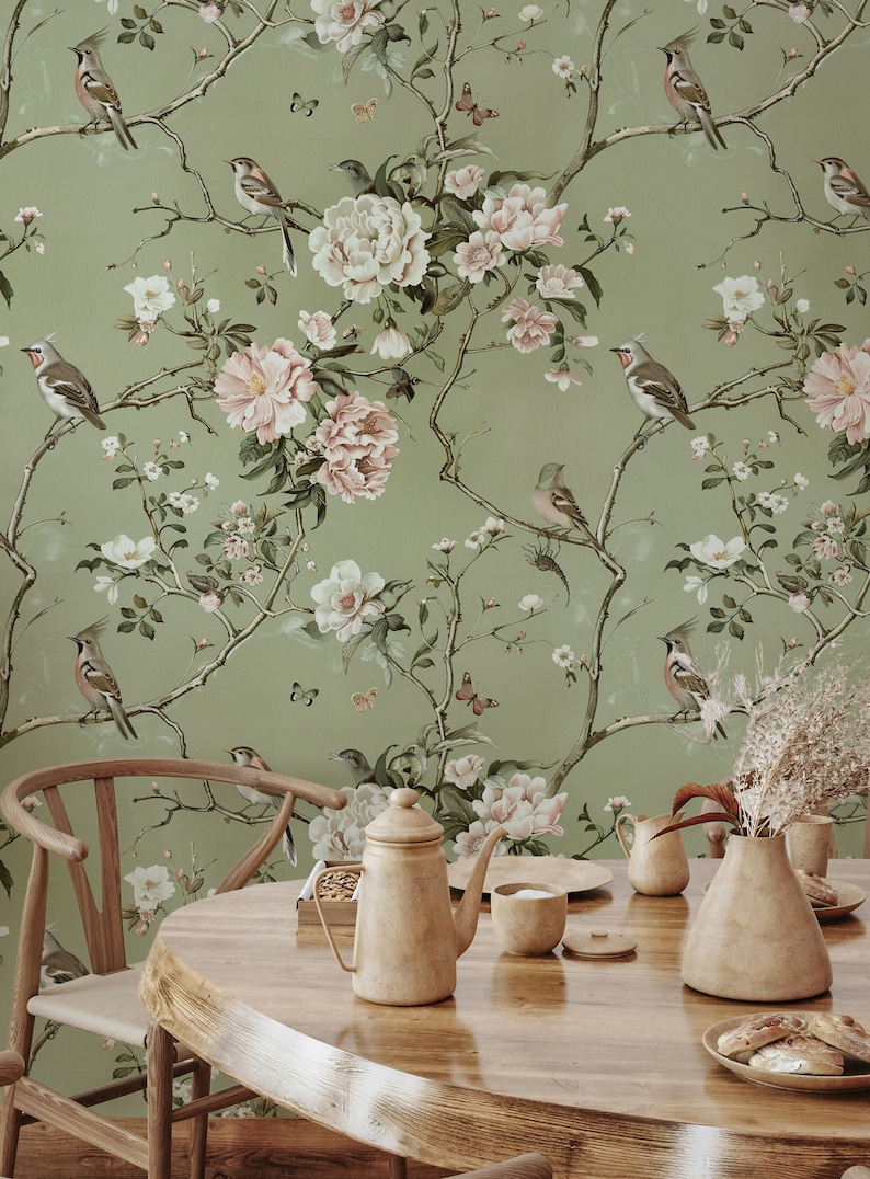 Op de afbeelding: Een groene behang met een bloemenpatroon met roze en witte bloemen, groene bladeren en vogels. Het behang bevindt zich in een eetkameromgeving met een houten tafel, stoelen en een theepot.