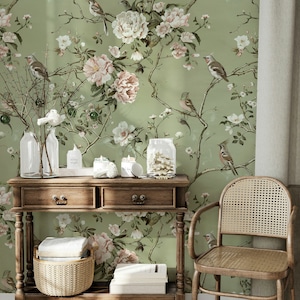 Op de afbeelding: Een houten consoletafel met twee laden en een onderplank. De tafel is versierd met een geweven mand en boeken. De tafel staat voor een muur met een groene bloemenbehang met roze en witte bloemen en vogels.