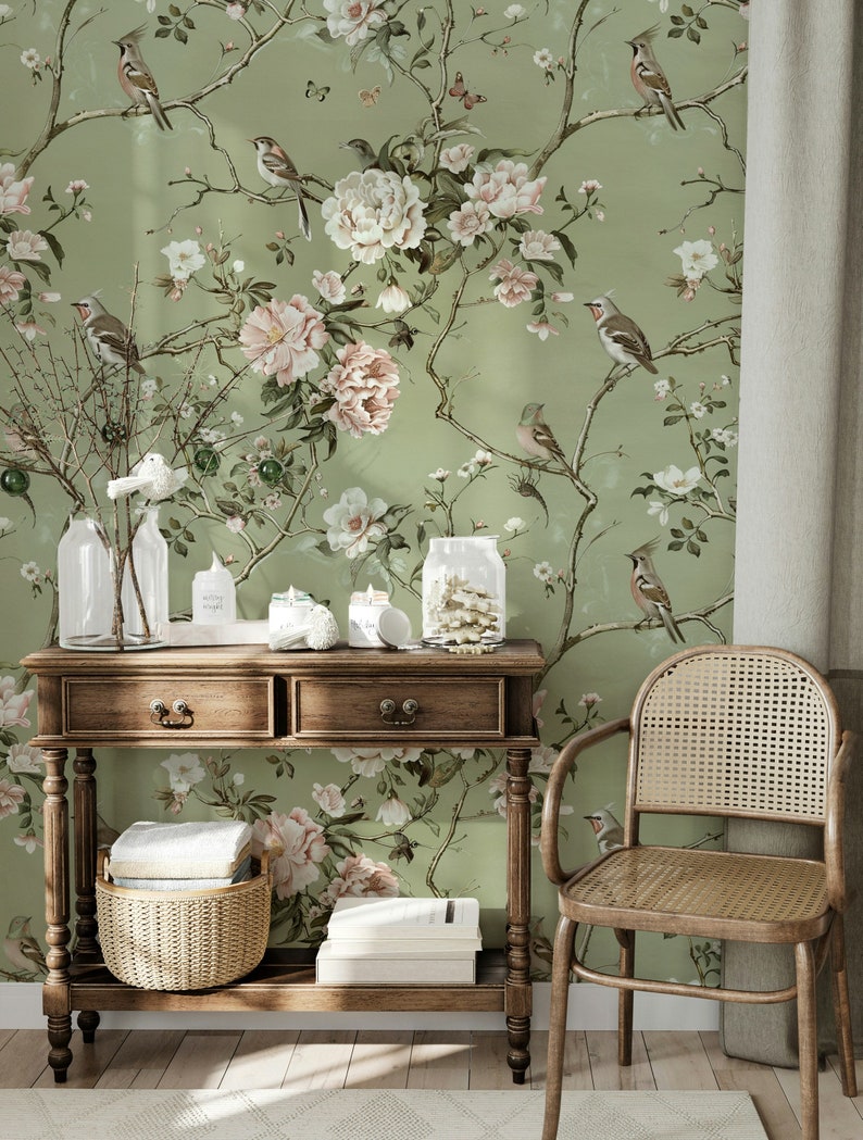 Op de afbeelding: Een houten consoletafel met twee laden en een onderplank. De tafel is versierd met een geweven mand en boeken. De tafel staat voor een muur met een groene bloemenbehang met roze en witte bloemen en vogels.