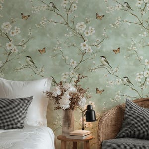 May include: A bedroom scene with floral wallpaper depicting birds and butterflies. A bed with white bedding and a patterned cushion is on the left. A wicker chair and a small wooden table with a lamp and books are on the right.