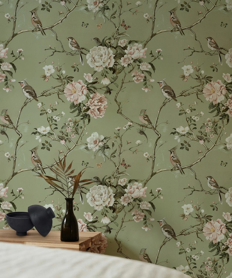 Op de afbeelding: Een groene behang met een bloemen- en vogelpatroon. Het behang heeft witte en roze bloemen, groene bladeren en bruine vogels. Het behang is te zien in een kamer met een houten tafel en een vaas met een plant.