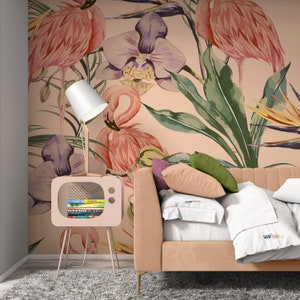 May include: A pink and green tropical wallpaper with pink flamingos and purple orchids. The wallpaper is behind a pink couch with white pillows and a pink bedside table with a lamp.
