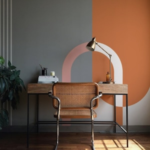Op de afbeelding: Een thuiskantoor bureau met een houten blad en zwarte metalen poten. Een stoel met een geweven rieten zitting staat voor het bureau. De muur achter het bureau is geschilderd in tinten grijs en oranje met een roze en wit geometrisch ontwerp.