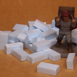 Könnte beinhalten: Eine Miniaturfigur eines Reisenden mit roter Kapuze und einem großen braunen Rucksack, die neben einem verstreuten Haufen kleiner, rechteckiger, hellblauer Schaumstoffblöcke steht, die wie Bausteine oder Baumaterialien aussehen.