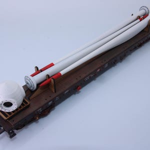 May include: A model of a train car carrying components of a wind turbine. The cargo includes three white and red blades and a white nacelle. The train car is brown and sits on a light blue surface.