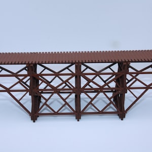 Könnte beinhalten: Ein braunes Holzmodell einer Bockbrücke. Die Brücke hat eine Reihe von abgewinkelten Stützen und ein Lattenrost. Das Modell ist für den Einsatz in Modelleisenbahnanlagen oder Dioramen konzipiert.