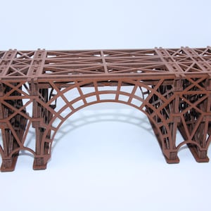 Può includere: Un modello tridimensionale di ponte marrone caratterizzato da un intricato design a traliccio. La struttura include una campata centrale ad arco sostenuta da due sezioni di pilastri rettangolari, il tutto costruito con una cornice geometrica ripetitiva.