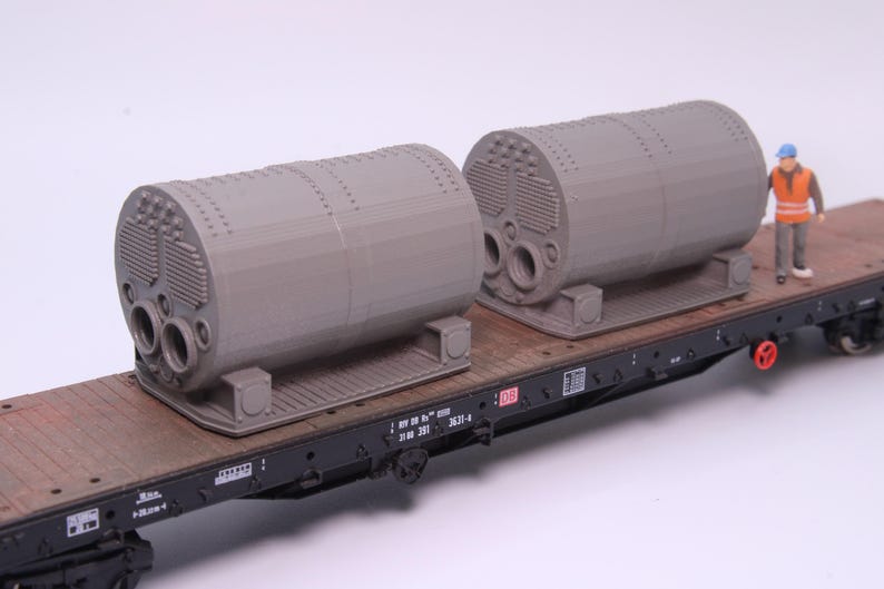 May include: A model train flatcar with a brown wooden deck carrying two grey cylindrical industrial boilers. Each boiler features a detailed front panel with circular openings and a base. A small scale figure wearing an orange vest stands nearby.