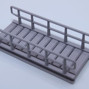 Könnte beinhalten: Ein kleines, graues Modell einer Brücke mit einem Lattenrost und Geländern. Die Brücke hat eine rechteckige Form und ist mit zwei parallelen Handläufen auf beiden Seiten versehen. Das Design ist einfach und funktional.