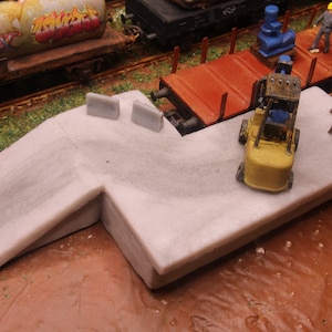 May include: A model railway scene featuring a light grey concrete platform and a yellow forklift. The miniature layout includes figures of workers, train carriages, and other equipment, creating a detailed diorama.