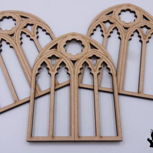 Może przedstawiać: Trzy wycięte laserowo drewniane kształty gotyckich okien. Okna mają jasnobrązowy kolor z czarnymi akcentami. Projekt obejmuje łukowate zwieńczenia i szczegółowe maswerki. Okna są ułożone na białej powierzchni.