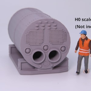 May include: A gray 3D printed model of a cylindrical steam locomotive boiler featuring a detailed front face with two large circular openings, smaller circular ports, and textured rivet patterns, accompanied by a small H0 scale figure.
