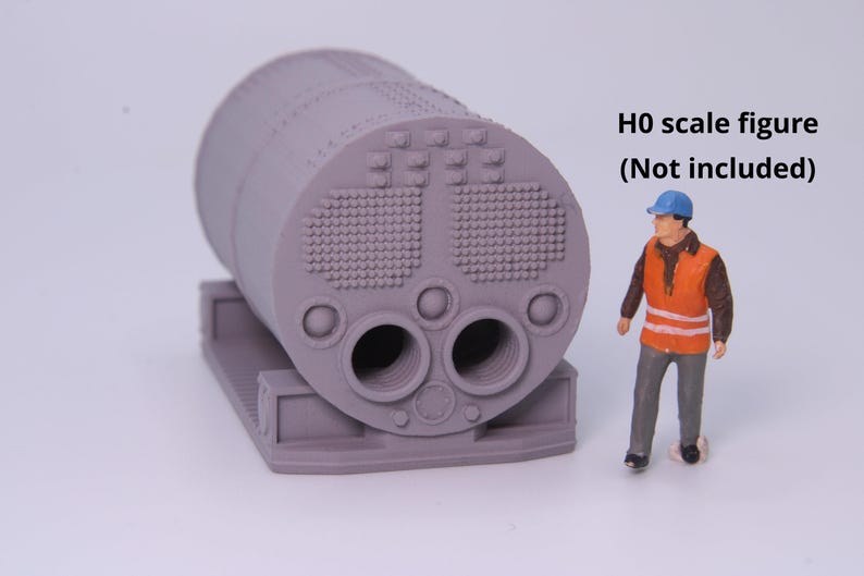 May include: A gray 3D printed model of a cylindrical steam locomotive boiler featuring a detailed front face with two large circular openings, smaller circular ports, and textured rivet patterns, accompanied by a small H0 scale figure.