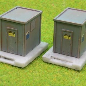 Pode incluir: Dois modelos cinzentos de edifícios utilitários com portas e aberturas, cada um montado numa base branca. As portas têm um sinal de aviso amarelo com um raio. Os edifícios estão sobre uma superfície verde.